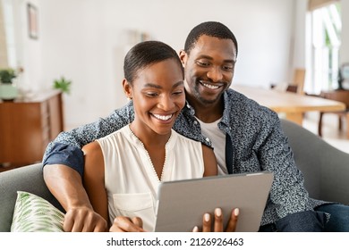 Cheerful black couple sitting on couch and watching movie on digital tablet at home. Mature black husband and beautiful wife using digital tablet to do a video call while sitting on sofa. - Powered by Shutterstock