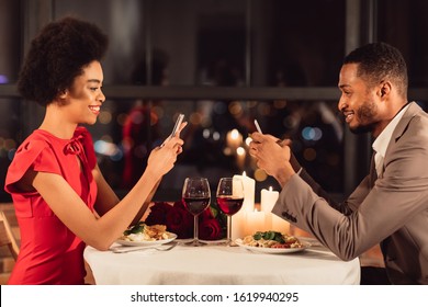 Cheerful Black Couple Using Cellphones Texting During Romantic Date Dinner In Fancy Restaurant. Modern Relationship And Lifestyle