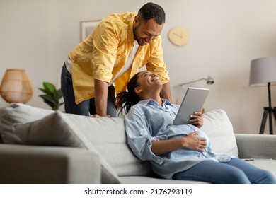 Cheerful Black Couple Using Digital Tablet Browsing Internet Sitting On Sofa At Home. Spouses Expecting Baby And Browsing Internet Via Computer On Weekend. Pregnancy Lifestyle, Technology