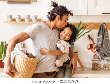 Cheerful Black Dad With Laundry Basket Gently Kisses Her Son  And Hugging Optimistic Kid While Doing Household Chores In Light Kitchen At Home