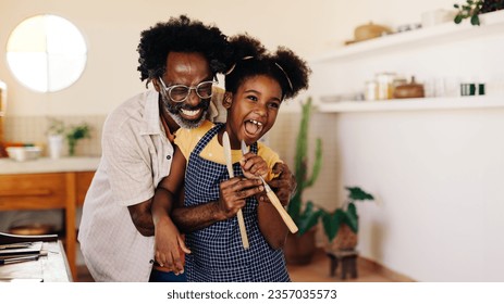 Cheerful black family in the kitchen. Father and daughter sing and smile while cooking, holding wooden spoons. Afro-haired dad wearing apron. Lively family time at home. - Powered by Shutterstock