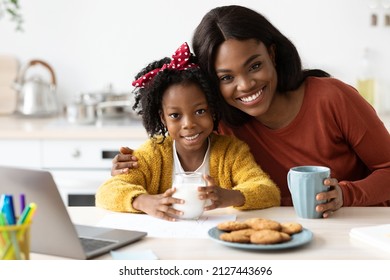 Cheerful Black Family Mom And Little Daughter Eating Snacks In Kitchen Together, Portrait Of Young African American Woman And Her Cute Female Child Enjoying Homemade Cookies And Drinking Milk