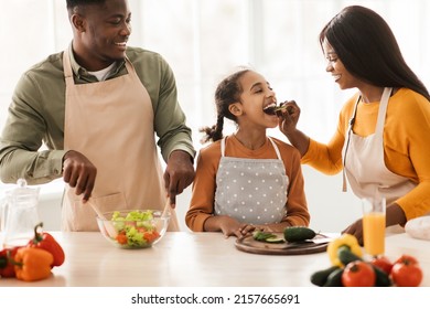 Cheerful Black Family Of Three Cooking Preparing Dinner, Mother Feeding Daughter While Making Fresh Vegetable Salad Standing In Modern Kitchen At Home. Healthy Food Recipes, Nutrition Concept