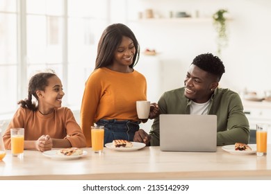 Cheerful Black Family Using Laptop Computer While Having Lunch Sitting At Table In Kitchen At Home. Joyful Parents And Daughter Eating Together And Browsing Internet Enjoying Domestic Weekend