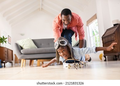 Cheerful black father pushing son lying on skateboard at home. Playful mature dad and son enjoying with skateboard and aviator cap in living room. Happy african american man with funny child playing.