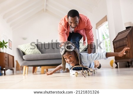 Cheerful black father pushing son lying on skateboard at home. Playful mature dad and son enjoying with skateboard and aviator cap in living room. Happy african american man with funny child playing.