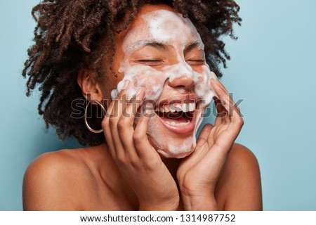 Cheerful black female model applies foaming cleanser, has clean fresh healthy skin, smiles broadly, Afro bushy haircut, stands bare shoulder against blue background. Beauty and feminine concept