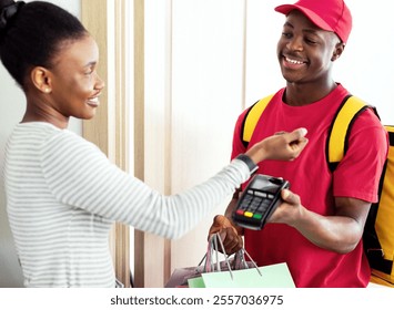 Cheerful Black Female Paying Courier Putting Smartwatch To Terminal Receiving Shopper Bags Standing At Home. Modern Shopping And Delivery Service. Selective Focus - Powered by Shutterstock