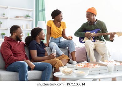 Cheerful Black Friends Playing Guitar And Singing Songs While Having Party At Home, Sitting On Couch In Living Room, Drinking Beer And Eating Pizza, African American People Relaxing At Weekend