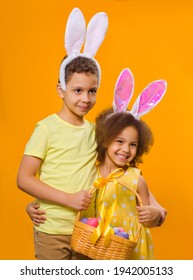 A Cheerful Black Girl And Boy With Rabbit Ears On Her Head With A Basket Of Colored Eggs In Her Hands On A Yellow Background. They Looks Away. Funny Happy Baby. Covid Easter To The Child.