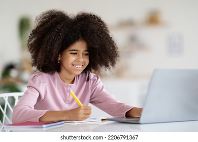 Cheerful black girl with bushy hair schooler sitting at desk at home, doing homework, using laptop, holding pen, taking notes, copy space. E-education for kids, home-schooling concept - Shutterstock ID 2206791367