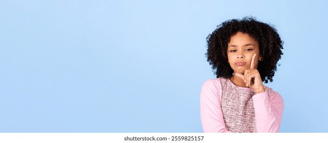 A cheerful black girl with curly hair poses while playfully resting her finger on her cheek. She showcases a moment of contemplation, surrounded by a light blue background, copy space - Powered by Shutterstock