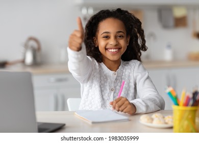 Cheerful black girl studying online, using laptop at home, taking notes or drawing, showing thumb up. Cute african american kid having online lesson, kitchen interior. Home schooling concept