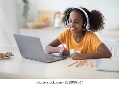 Cheerful black girl studying online, using laptop and headset at home, copy space. Cute african american teen kid having online lesson, drawing, kitchen interior. Home schooling concept