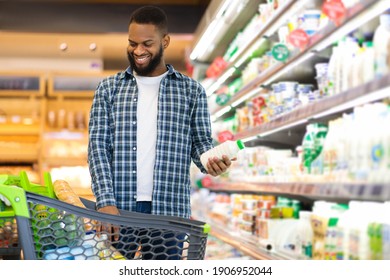 Cheerful Black Guy Doing Shopping Buying Groceries In Supermarket Standing With Cart Full Of Food, Choosing Milk At Shelf With Dairy Products Indoors. Copy Space