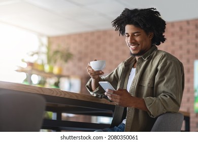 Cheerful Black Guy Drinking Coffee And Using Smartphone While Sitting At Table In Cafe, Happy Young African American Man Browsing Internet On Cellphone Or Reading Morning News Online, Copy Space