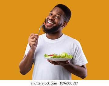 Cheerful Black Guy Eating Salad Posing Holding Plate And Fork Enjoying Healthy Meal Standing Over Yellow Studio Background, Smiling To Camera. Vegan Dinner Recipes, Weight Loss And Male Nutrition