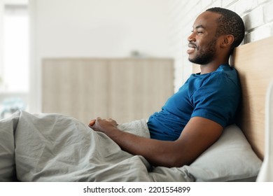 Cheerful Black Guy Sitting In Cozy Bed Smiling Looking Aside Posing In Modern Bedroom At Home. Good Morning, Healthy Sleeping Routine Concept. Side View Shot