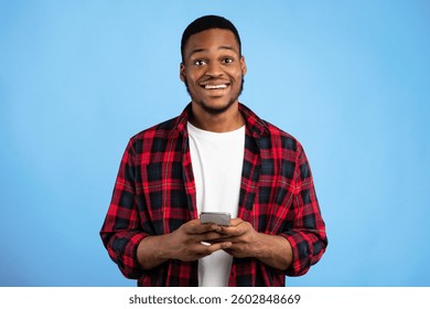 Cheerful Black Guy Using Smartphone Smiling To Camera Standing Over Blue Background. Studio Shot