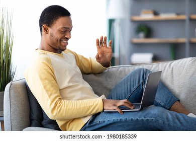 Cheerful Black Guy Waving At Laptop Screen, Reclining On Couch And Having Video Call With Friends Or Lover, Home Interior, Side View, Copy Space. Communication During COVID-19 Pandemic