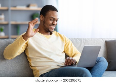 Cheerful Black Guy Waving At Laptop Screen, Reclining On Couch And Having Video Call With Friends Or Lover, Home Interior, Side View, Copy Space. Communication During COVID-19 Pandemic