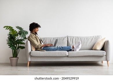 Cheerful Black Guy Working On Laptop While Sitting On Couch At Home, Happy Young African American Male Freelancer Typing On Keyboard, Enjoying Remote Work And Distance Communication, Side View