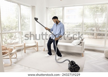 Cheerful Black homeowner guy having fun while tidying up at home, vacuuming carpeted floor in living room, dancing to music, singing song, imitating guitar playing with hoover