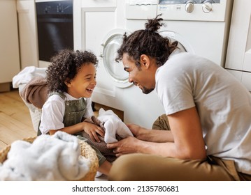 Cheerful Black Kid Boy Helping Father At Linen In Basket While Doing Laundry Near Washing Machine In Flight Kitchen In Weekend At Home  