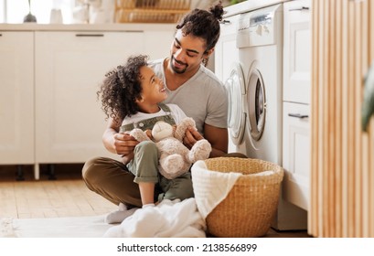 Cheerful black kid boy helping father at linen in basket while doing laundry near washing machine in flight kitchen in weekend at home  