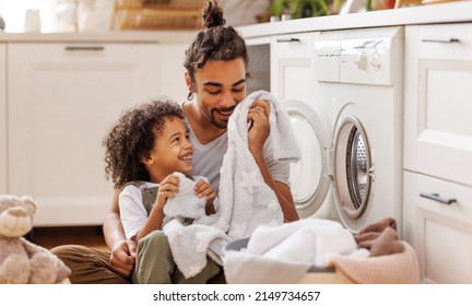 Cheerful Black Kid Boy Helping Father At Linen In Basket While Doing Laundry Near Washing Machine In Flight Kitchen In Weekend At Home  