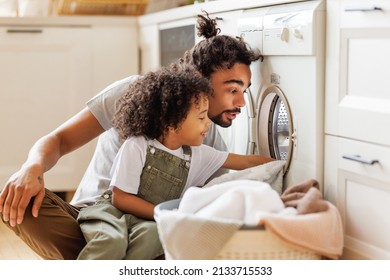 Cheerful black kid boy sitting on dad's lap and helping father at linen in basket while doing laundry near washing machine in flight kitchen in weekend at home   - Shutterstock ID 2133715533