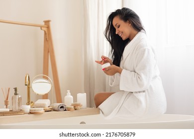 Cheerful Black Lady Applying Moisturizer Holding Opened Jar Moisturizing Skin And Caring For Body Sitting On Bathtub In Modern Bathroom Indoor. Beauty Care And Cosmetics