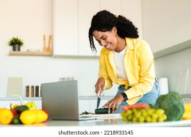 Cheerful Black Lady Cutting Organic Vegetables And Using Laptop, Cooking Food In Minimalist Kitchen Interior, Free Space. Technology, Blog, New Salad Recipe, Veganism And Health Care