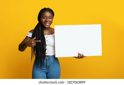 Cheerful Black Lady Holding And Pointing At White Blank Placard, Demonstrating Free Place For Your Text Or Advertisement, Showing Copy Space On Billboard, Posing Over Yellow Studio Background