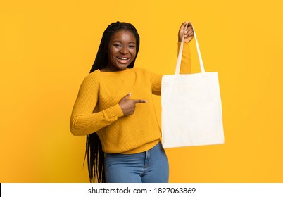 Cheerful Black Lady Pointing At Blank Textile Tote Eco Canvas Shopping Bag In Her Hand With Free Copy Space For Your Design Or Logo, Looking At Camera While Posing Over Yellow Studio Background