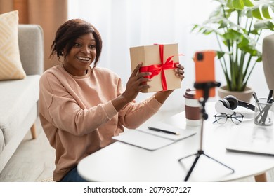 Cheerful Black Lady Showing Christmas Gift To Phone During Video Call Celebrating Holiday Online Or Making Video For Blog At Home. Modern Blogging Concept. Selective Focus