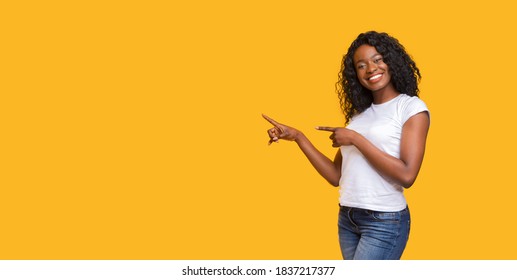 Cheerful black lady in white t-shirt pointing at copy space on yellow studio background. Smiling african american young woman showing exciting advertisement or announcement, panorama with empty space