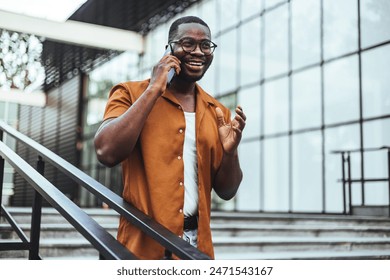 A cheerful black male student talks on his phone while ascending stairs outside a campus building, dressed in casual attire with eyeglasses. - Powered by Shutterstock