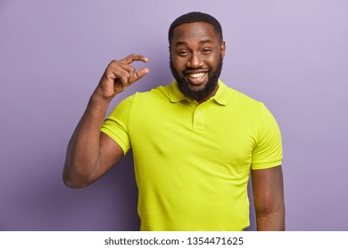 Cheerful Black Man With Bright Smile, Shows White Teeth, Shapes Something Very Tiny With Hand, Wears Yellow T Shirt, Stands Over Purple Background, Pretends Holding Small Object, Isolated On Lilac