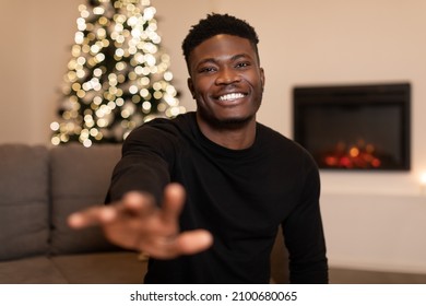 Cheerful Black Man Stretching Hand To Camera And Smiling Sitting On Couch Celebrating New Year At Home. Modern Remote Holiday Celebration Concept. Selective Focus