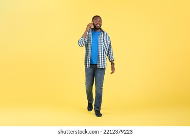 Cheerful Black Man Talking On Cellphone Posing Walking Smiling To Camera On Yellow Background In Studio. Mobile Communication And Gadgets Concept. Full Length Shot