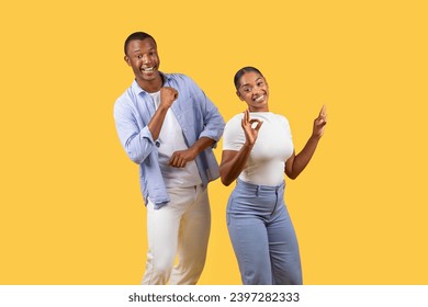 Cheerful black man and woman making positive hand gestures, dancing and woman signaling OK, both radiating happiness against yellow backdrop