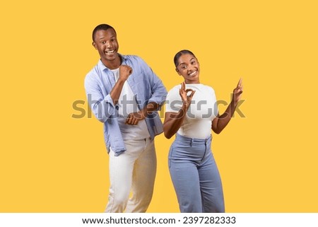 Cheerful black man and woman making positive hand gestures, dancing and woman signaling OK, both radiating happiness against yellow backdrop