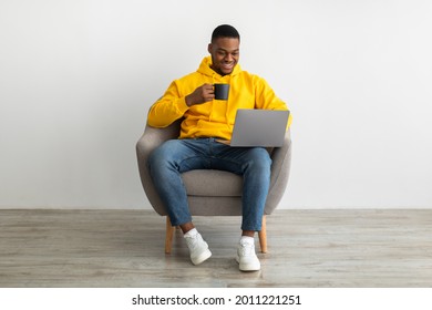 Cheerful Black Man Working On Laptop Computer Online Drinking Coffee Sitting In Armchair Over Gray Wall Indoors. Guy Browsing Internet At Home. Technology, People And Gadgets Concept