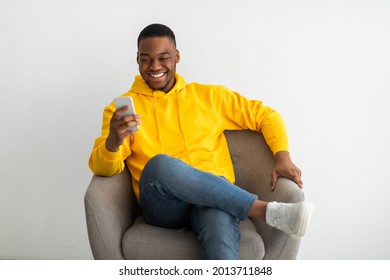 Cheerful Black Millennial Man Using Cellphone Browsing Internet And Texting Sitting In Armchair Over Gray Wall Indoors. People And Gadgets. Mobile Communication Concept