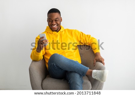 Cheerful Black Millennial Man Using Cellphone Browsing Internet And Texting Sitting In Armchair Over Gray Wall Indoors. People And Gadgets. Mobile Communication Concept Foto stock © 