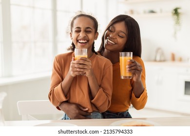 Cheerful Black Mommy And Daughter Drinking Orange Juice And Laughing Holding Glasses Sitting Embracing In Modern Kitchen Indoors. Family Nutrition, Healthy Eating Concept