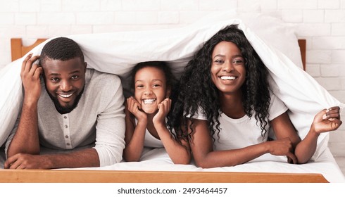 Cheerful black mother, father and daughter hiding under blanket, playing on bed at home, copy space - Powered by Shutterstock