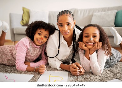 Cheerful Black Mother Of Two Preteen Daughters Drawing And Having Fun With Kids With Painted Faces Posing Smiling To Camera Lying On Floor At Home  Family Weekend Activity And Hobby