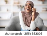 Cheerful black muslim lady in hijab talking on cellphone at home, relaxing on comfortable chair in living room, happy religious african woman having pleasant conversation with friend, copy space